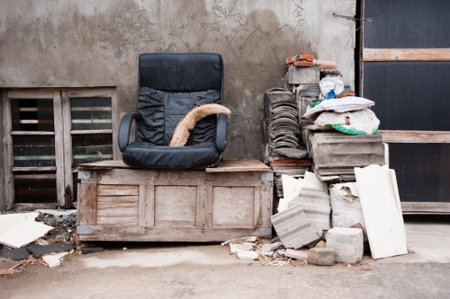 The image depicts an outdoor scene with a worn, black office chair positioned on a wooden crate or pallet against an aged, weathered concrete wall. To the right of the chair, there is a pile of construction and renovation debris, including stacked bricks, concrete blocks, and various pieces of broken or discarded building materials. Several plastic bags filled with waste are also visible, resting on top of the brick stack. The setting appears to be an alleyway or a part of a back alley near residential or commercial premises, with a vent opening near the ground on the left side of the image and a black metal gate or door on the right. The ground surface is concrete, showing signs of dirt and small debris, characteristic of a typical rubbish disposal or clearance area in Dalston, which could benefit from the services of Garden Clearance Dalston for debris removal and rubbish clearance.