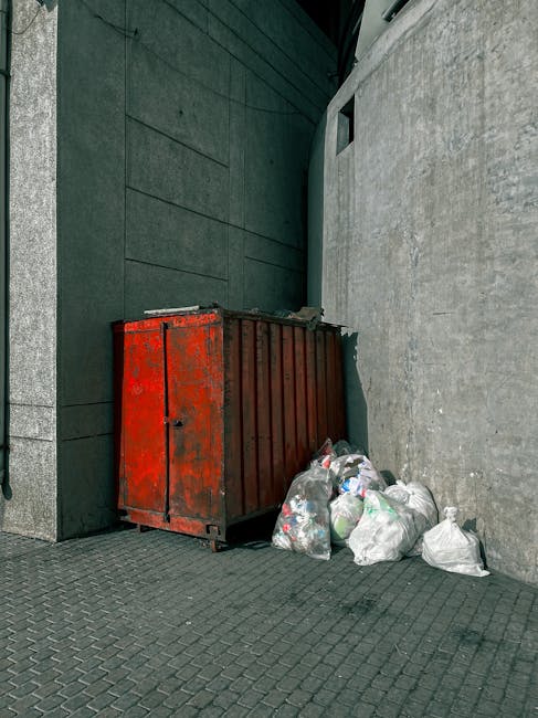 A man with dark hair, wearing a black T-shirt with white text, is seen leaning forward and placing a large white plastic bag filled with waste into a silver, cylindrical outdoor rubbish bin made of me